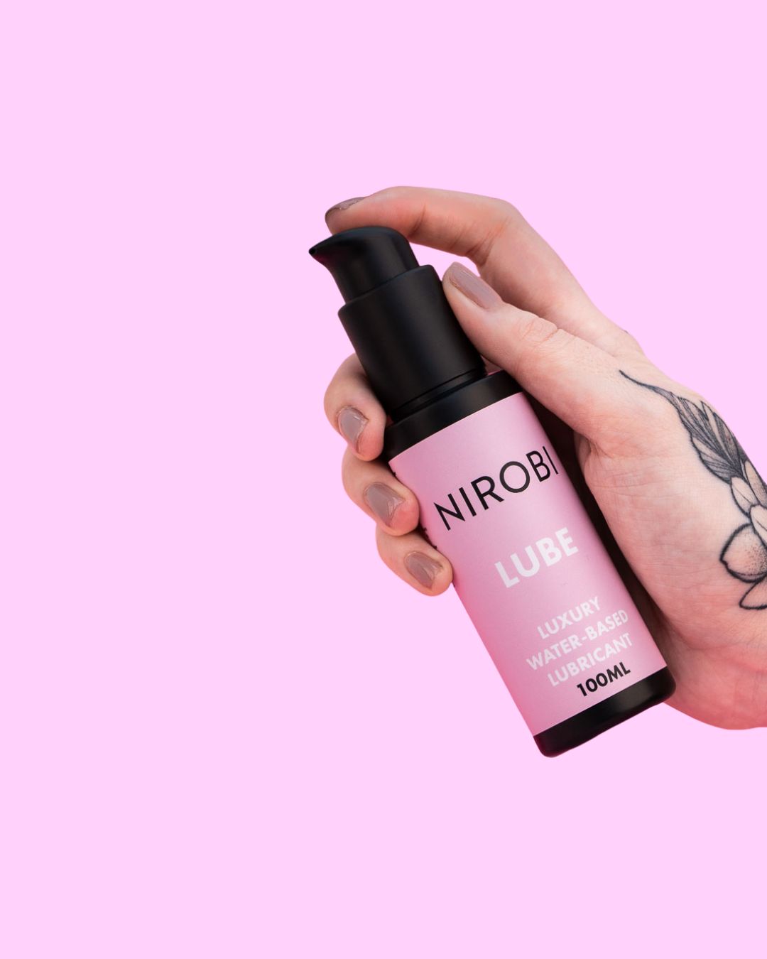 Close-up of a tattooed hand holding a Nirobi 100 ml water-based lubricant pump bottle on pink.
