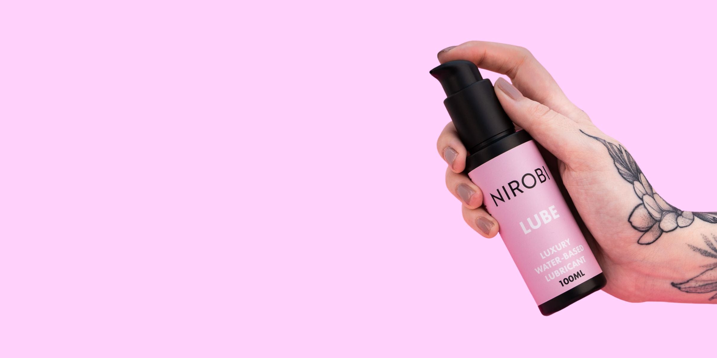 Landscape banner with a tattooed hand holding a Nirobi water-based lube bottle on a pale pink background.